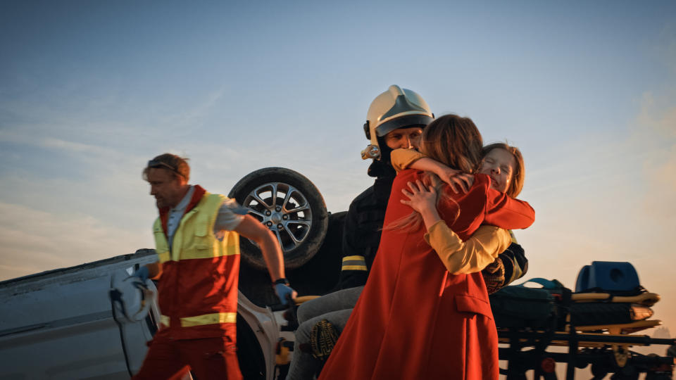 People embracing while standing in front of an upside-down car