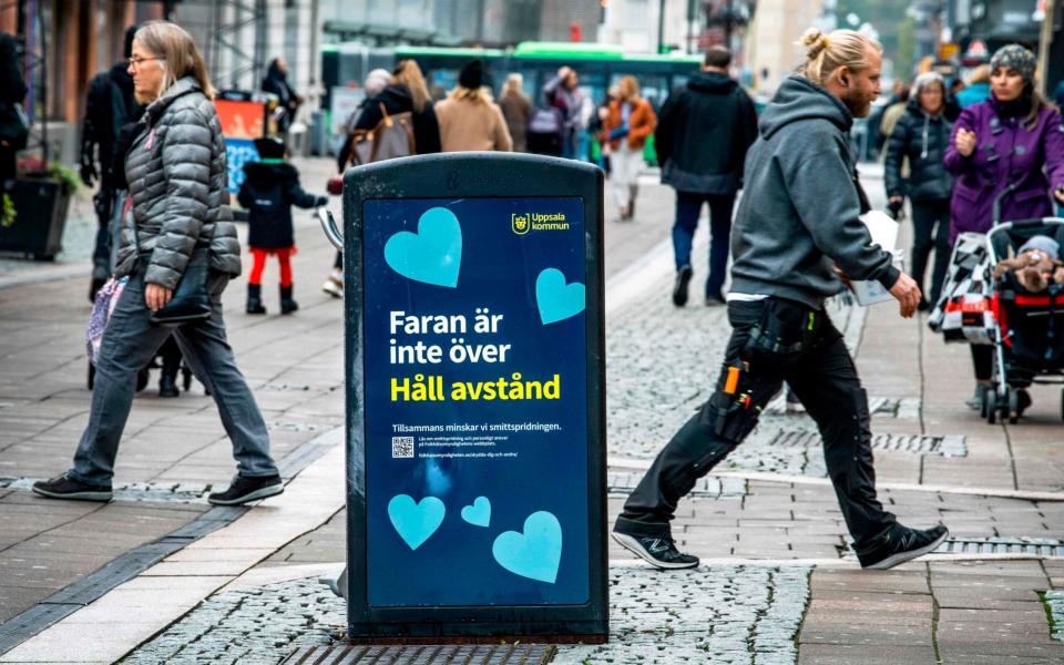 People walk past a bin with a sign reading "The danger is not over - Keep your distance" in a pedestrian street in central Uppsala, the first town in Sweden to enter local restrictions - CLAUDIO BRESCIANI /AFP