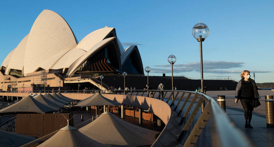 A woman is seen wearing a mask while walking in front of the Sydney Opera House