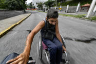 A man wearing a protective face mask hitches a ride in his wheelchair by grabbing on to the back of a motorcycle, near the Miraflores Palace in Caracas, Venezuela, Saturday, Aug. 8, 2020, amid the new coronavirus pandemic. (AP Photo/Matias Delacroix)