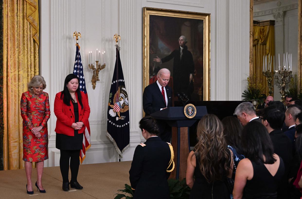 First lady Jill Biden, in red silk dress, Elaine Tso, in red jacket, and President Biden, facing a large audience in formal dress, bow their heads.
