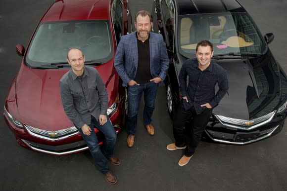 The three are standing with two Chevrolet Volt sedans.