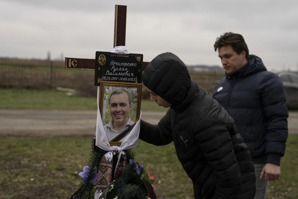 Yura Nechyporenko besa una foto de su padre, Ruslan Nechyporenko, junto a su tío Andriy Nechyporenko en el cementerio en Bucha, a las afueras de Kiev, Ucrania, el jueves 21 de abril de 2022. El joven sobrevivió a un intento de ejecución de soldados rusos, mientras que su padre murió, y ahora la familia pide justicia. (AP Foto/Petros Giannakouris)