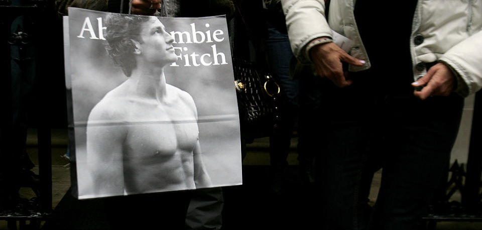 A person carries an Abercrombie & Fitch shopping bag featuring a black-and-white image of a shirtless male model. Another person is next to them