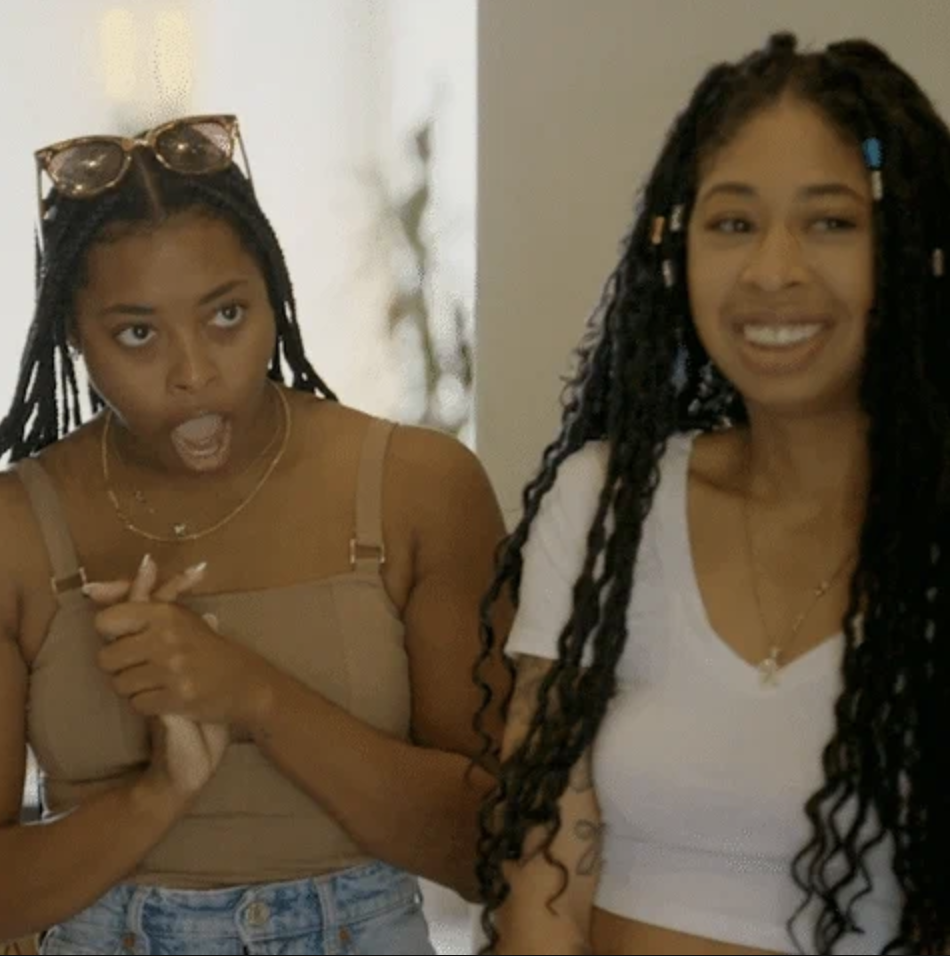 Two women with long braided hair look pleasantly surprised and smile in an indoor setting. One woman wears a tan top and the other a white top. Names not provided