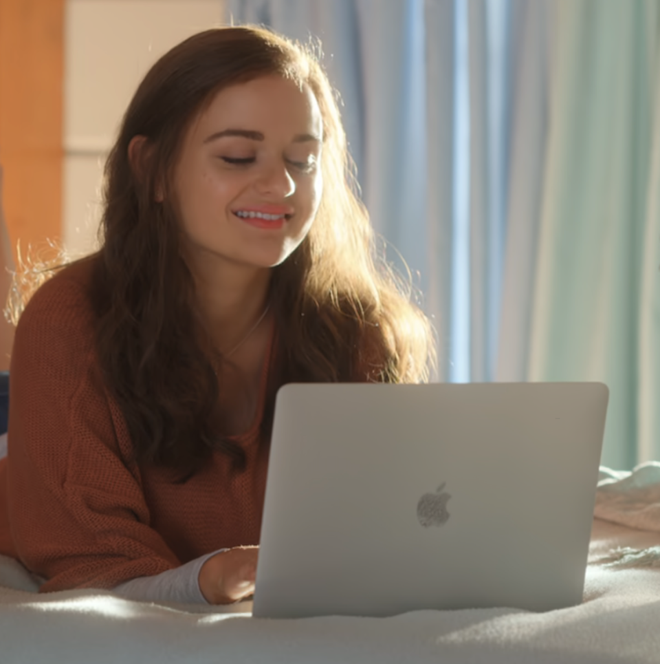 Joey King lying on bed, smiling at her laptop screen. She is wearing a casual long sleeve top. Curtains and window visible in the background