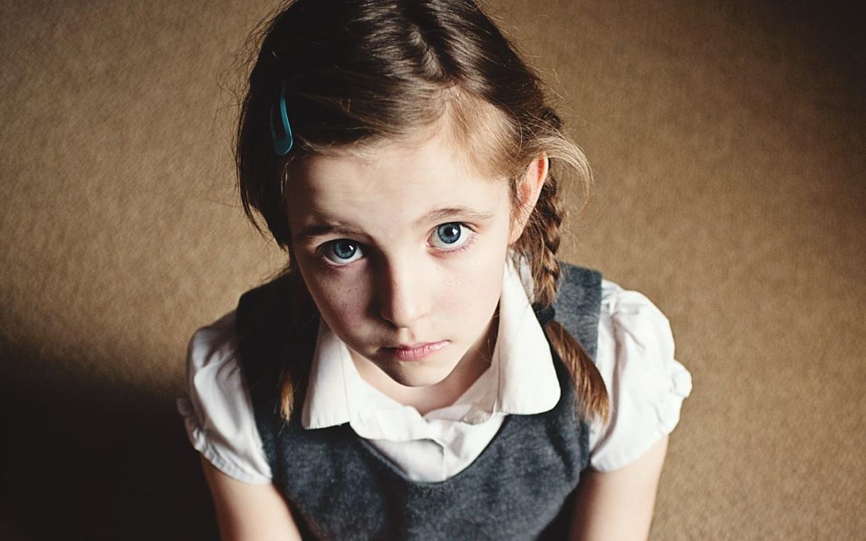 This is a picture of a sad/unhappy looking 7 year old girl. The photo is taken from above and she is sat cross legged on the floor. She is wearing school uniform with pink tights. She has a toy in her hand. She looks lonely. She has plaits in her hair. -  Getty Images/ Getty Images