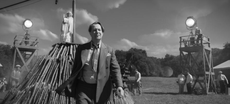 A woman in white watches from a platform as a man walks through an outdoor movie set.