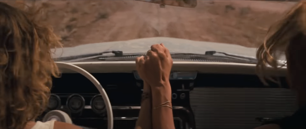Thelma and Louise from "Thelma and Louise" holding hands while driving on a dirt road in a convertible, the scene suggests a sense of adventure and love