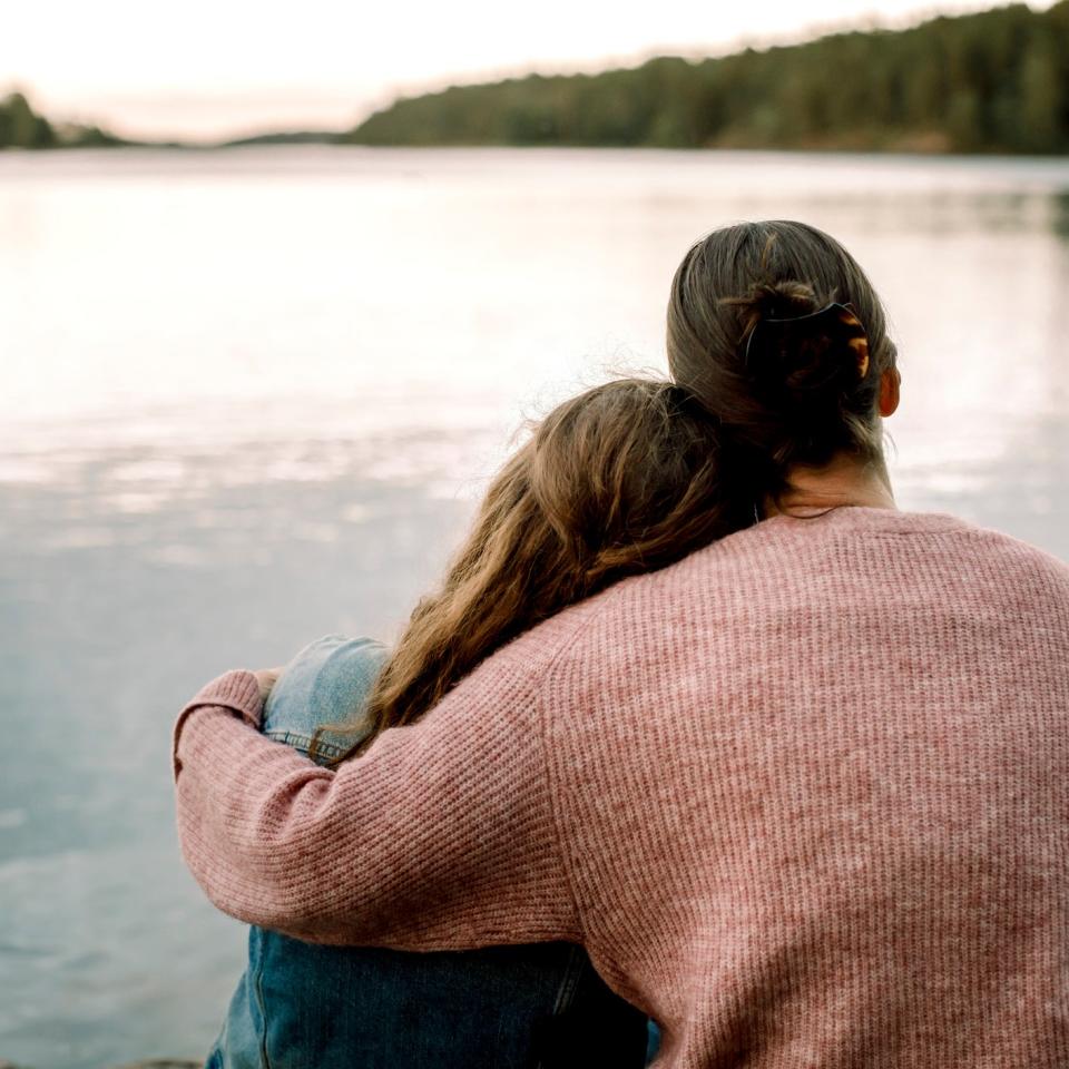 Two people sit by a serene lake, one with their arm around the other, conveying a sense of comfort and connection