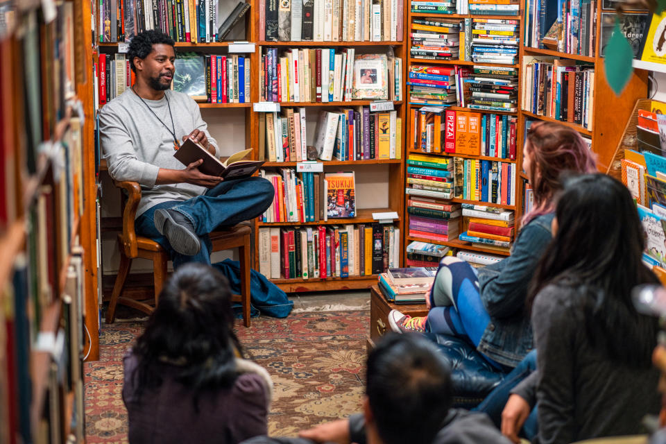 A person sits in a bookstore, reading a book to a small seated audience. Around them are shelves full of books.