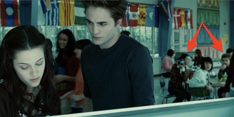 Bella and Edward talking in the lunch line with three of her friends sitting at a table in the background and staring at her