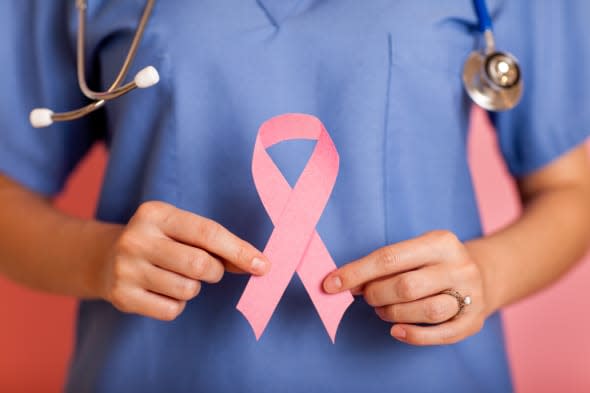 Color photo of a female nurse holding up a Breast Cancer Awareness Ribbon on pink background.