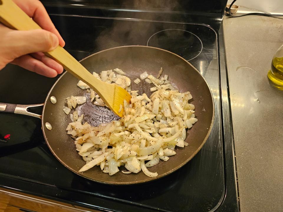 Hand holding wooden spoon and sauteeing onions in a pan on a stovetop