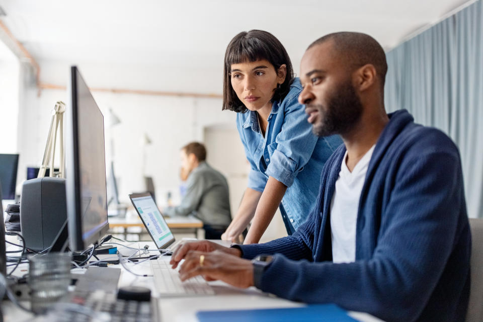 Two people work at computers in an office. One person sits and types while the other person stands and leans over to assist