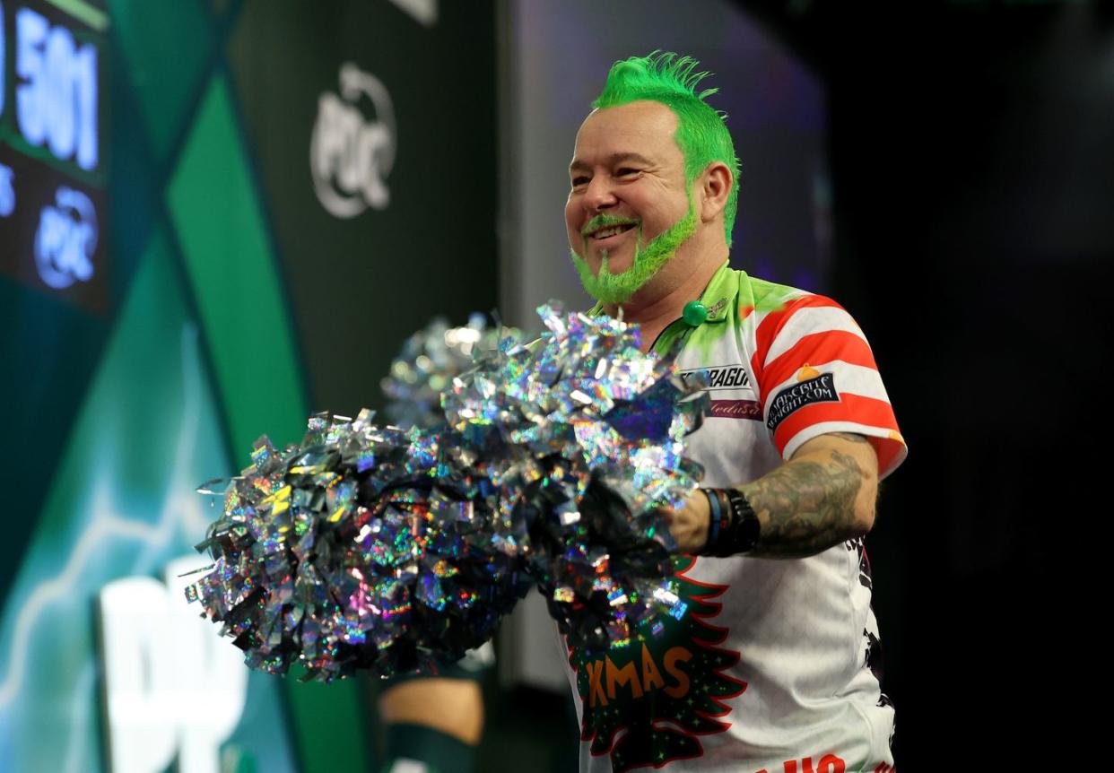 peter wright, a darts player with dyed green hair and beard, smiles and holds metallic pompoms in his hands as he entertains the crowds at a tournament