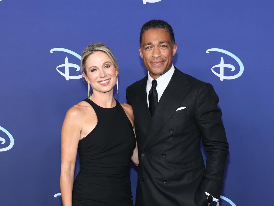 amy robach, wearing a blach dress with an asymmetric, ruffled hem, and t.j. holmes, wearing a black suit and black tie, standing together in front of a navy background with the Disney logo