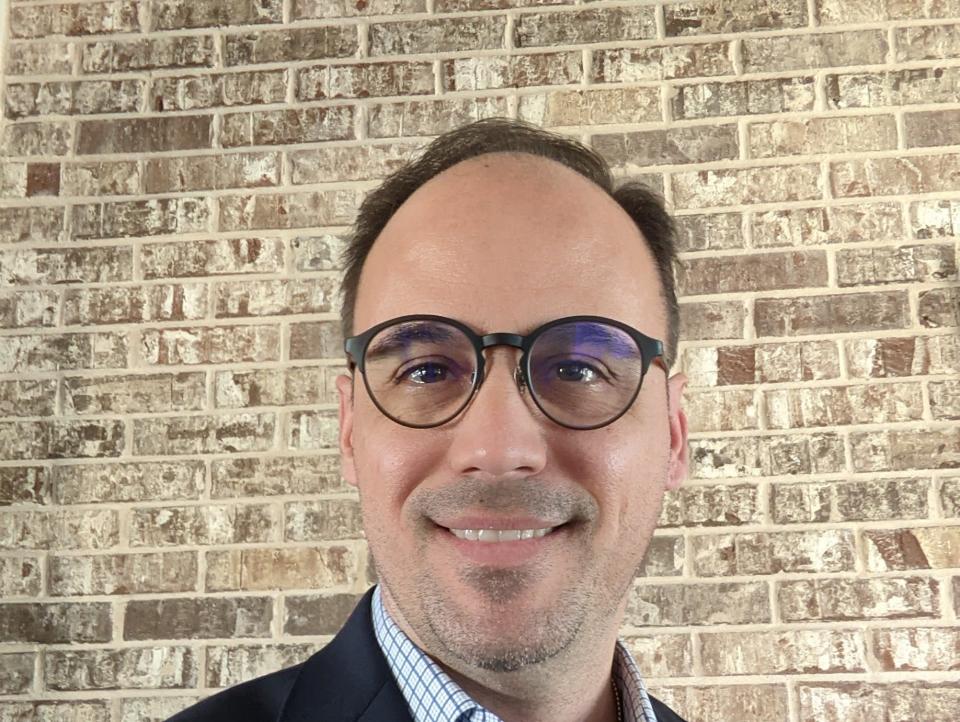 Tony Vargas poses near a brick wall in a check shirt, blue jacket and round glasses