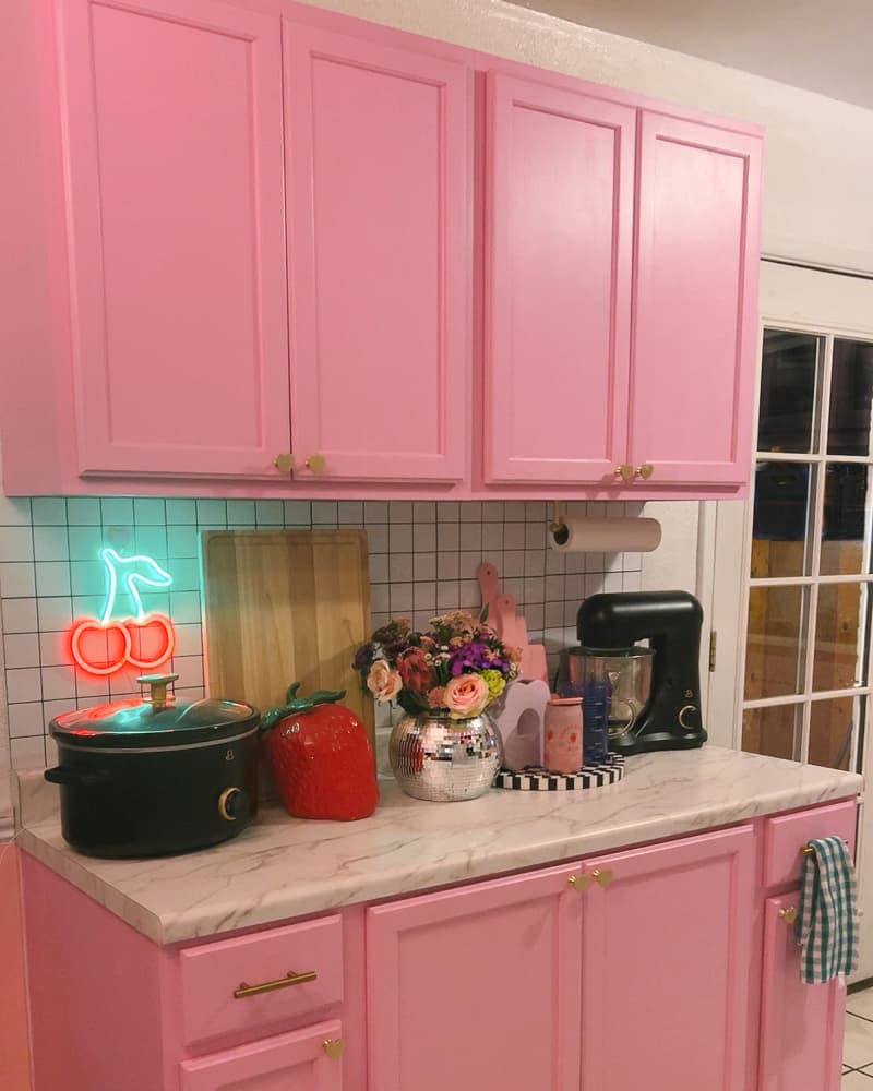 Pink cabinets with marble countertop holding small appliances and flowers in front of white tile backsplash with neon cherry sign.