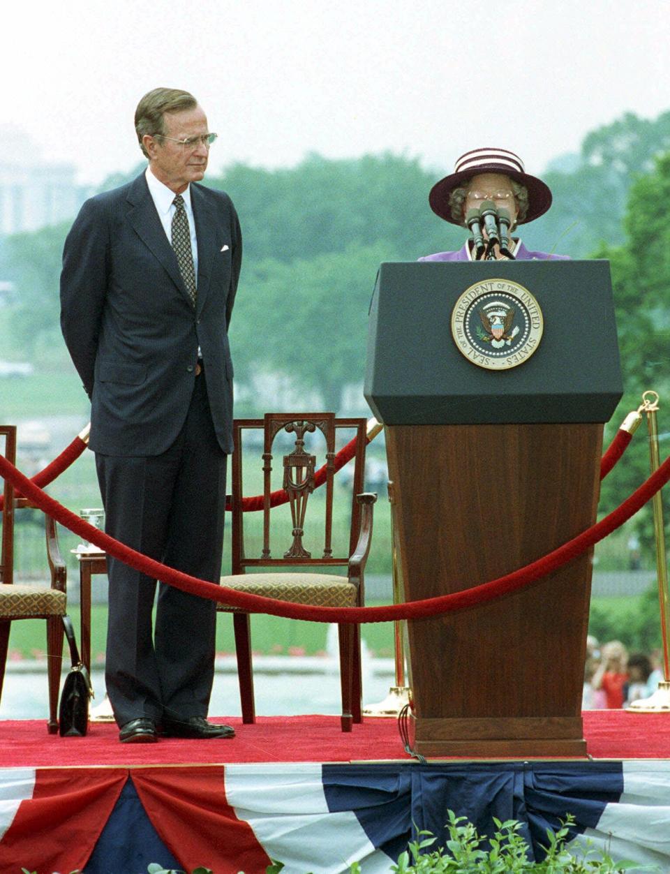 Queen Elizabeth II peers over microphones while giving arrival remarks on the White House South Lawn as President George H.W. Bush looks on May 14, 1991.