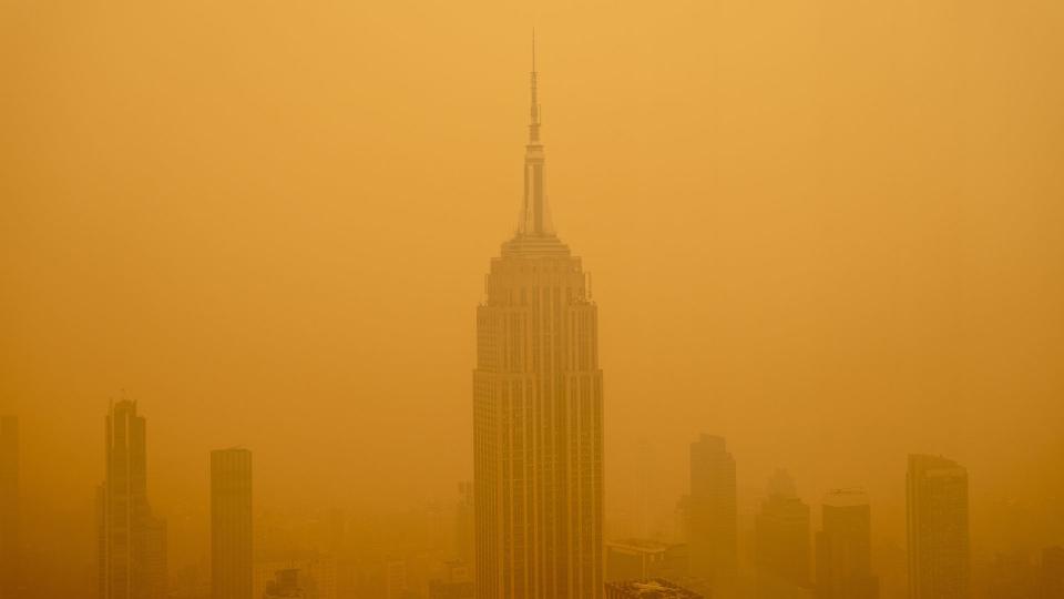 photo shows the empire state building as viewed from a nearby skyscraper; the building and those nearby are surrounded by a dense cloud of yellow-orange smog made up of wildfire smoke