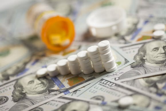 Ascending stacks of prescription tablets next to a tipped-over pill bottle, lying on a messy pile of hundred dollar bills.