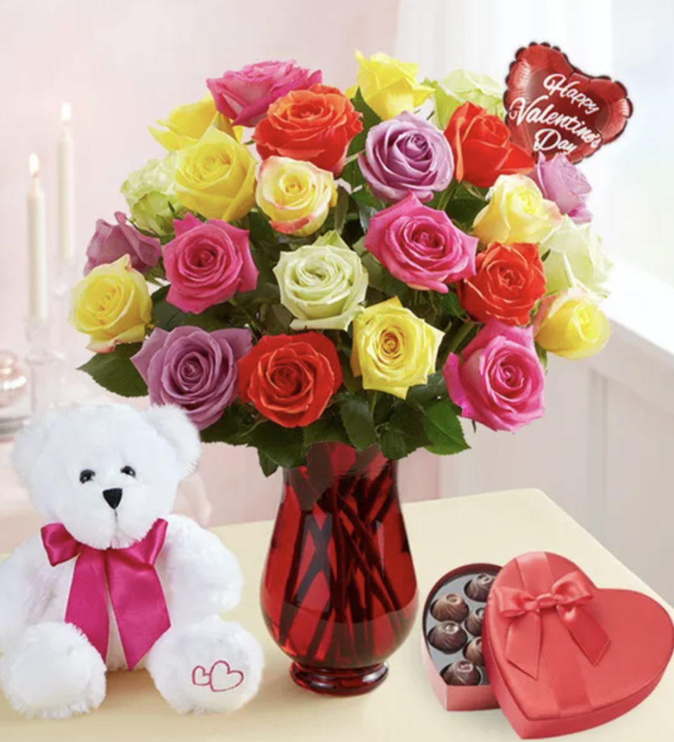 Red vase filled with pink, yellow, lavender, and red roses and a white teddy bear and heart shaped box of chocolates sitting nearby. 