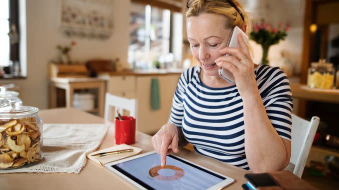 Beautiful senior woman at home working on tablet sitting at ther kitchen table, searching or reading something and making phone call.