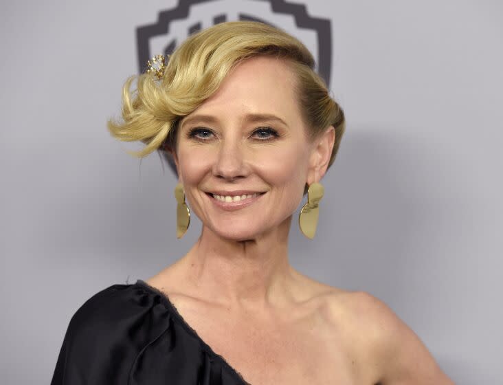 A woman with blond hair in an updo posing in gold earrings and an off-shoulder black dress