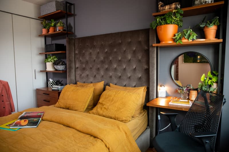 Bedroom. Bed with gold bedding, taupe quilted headboard, shelves with plants. Small desk with desk chair and mirror