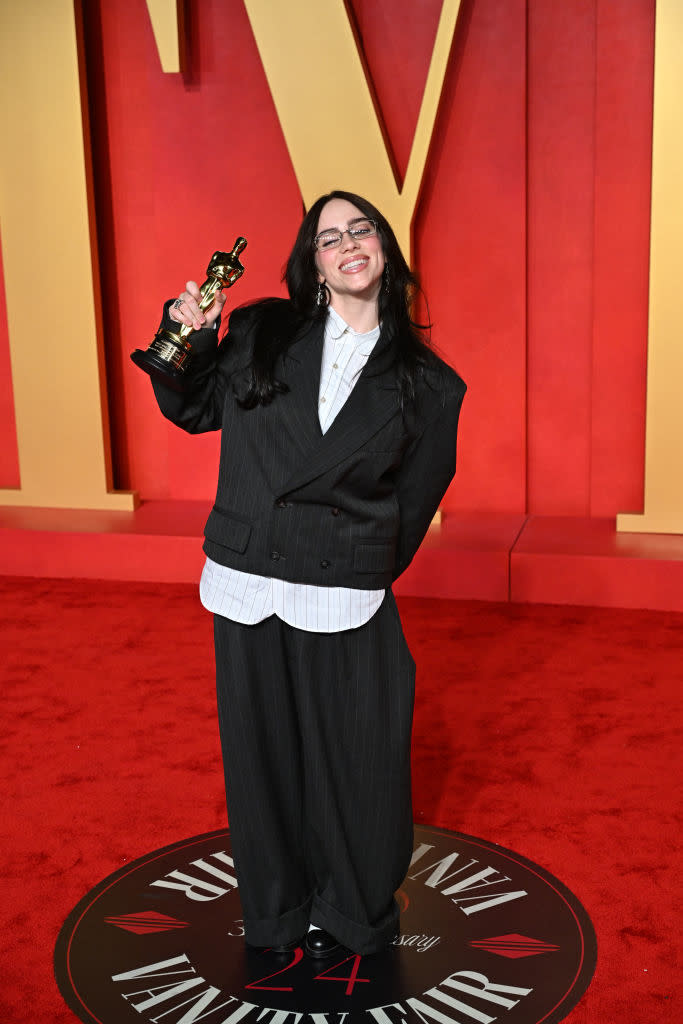 Person in pinstripe suit holding an award on a red carpet with a big letter "M" in the background