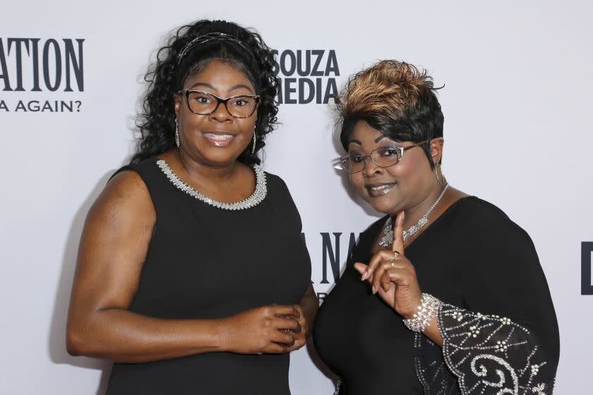 A woman with long, curly black hair and glasses and a woman with short, multicolored hair and glasses posing in black dresses