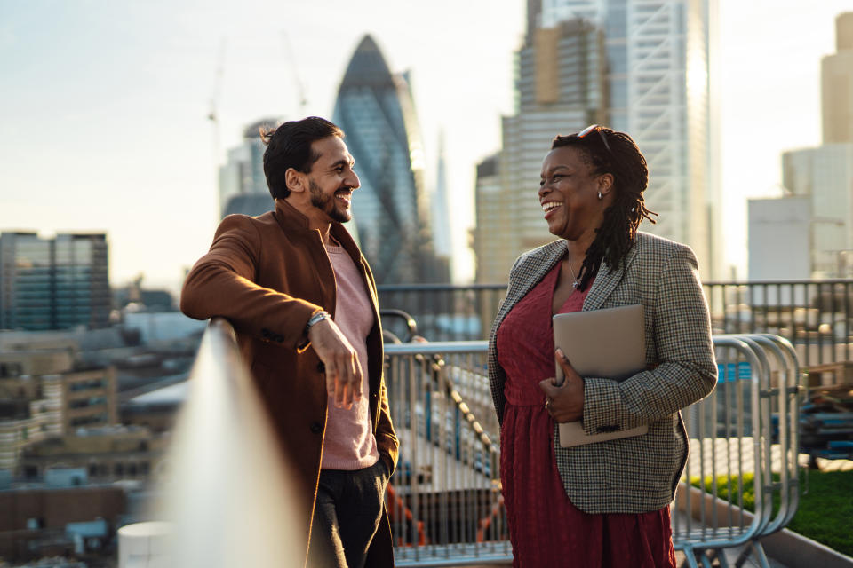 Mixed race businessman talking with an African businesswoman on city sidewalk. Multiracial business team having work discussion at rooftop garden in a high-rise office building. They're talking, planning and thinking strategy and discussing business strategy at urban balcony overlooking city, Liverpool, London. Holding laptop.