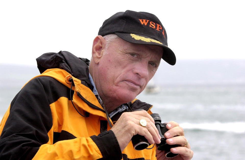 Ric O'Barry wears a black cap and holds binoculars. He is standing in front of the ocean.