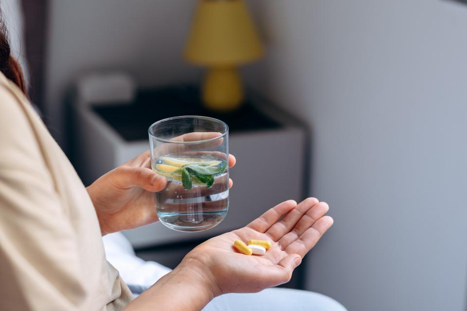 A person holds three capsules in one hand and a glass of water with lemon and mint in the other. 