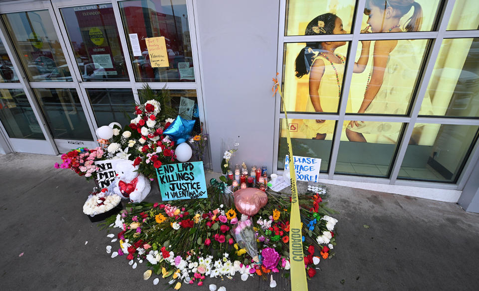 Flowers, signs and stuffed toys left outside a shop window with yellow caution tape on the side.