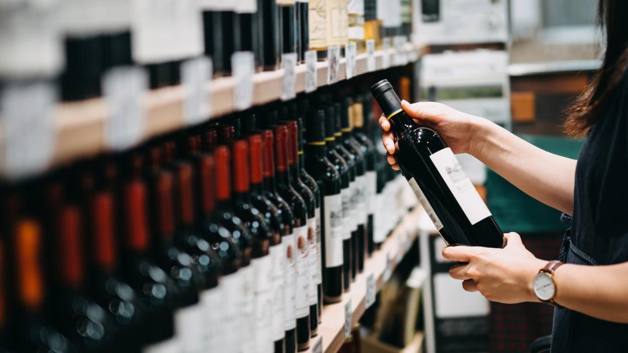 close up of young asian woman walking through supermarket aisle and choosing a bottle of red wine from the shelf in a supermarket