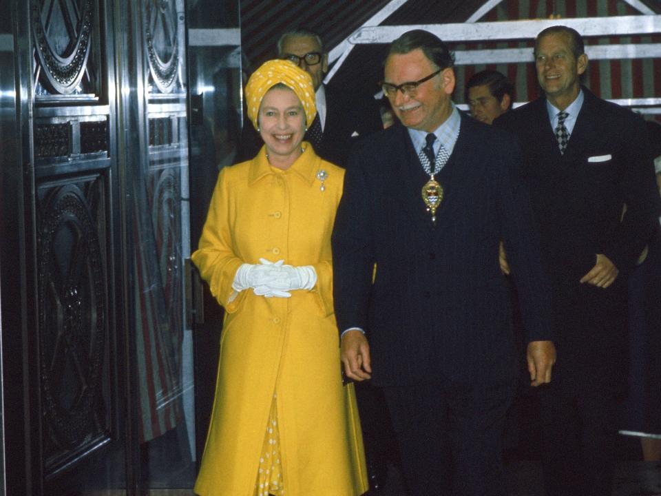 The queen smiling in a bright yellow overcoat with large buttons and a brooch and a polka-dotted dress underneath. She's wearing a yellow and white polka dot turban on her head and white gloves.