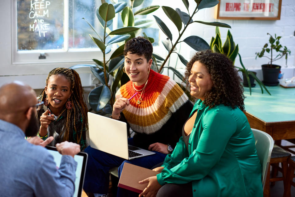 Four people in a casual office meeting, one with a laptop, engaging in a discussion