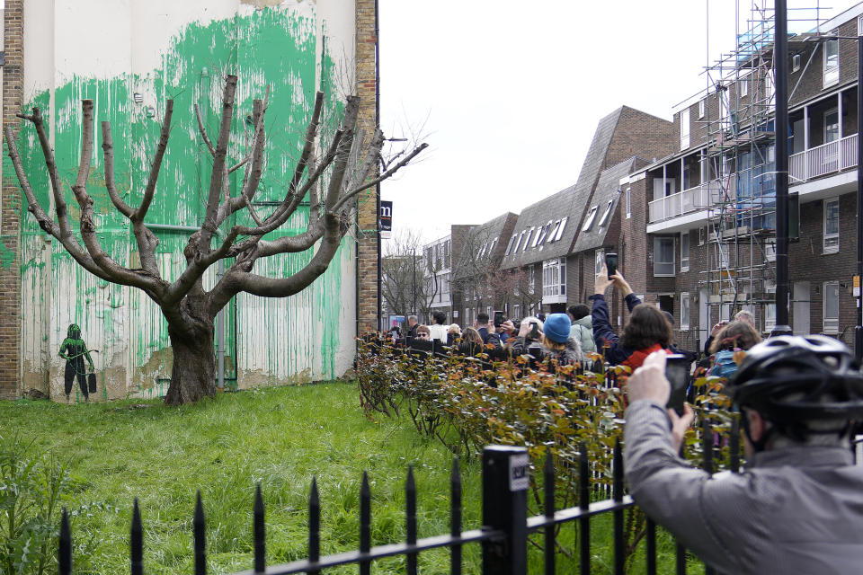 People look at a new Banksy painting on a wall in London, Monday, March 18, 2024. A new Banksy mural drew crowds to a London street on Monday, even before the elusive graffiti artist confirmed that the work was his. The artwork in the Finsbury Park neighborhood covers the wall of a four-story building and shows a small figure holding a pressure hose beside a real tree. Green paint has been sprayed across the wall, replicating the absent leaves of the tree, which has been severely cropped. Banksy claimed the work by posting before and after photos of the location on his official Instagram account.(AP Photo/Alastair Grant)