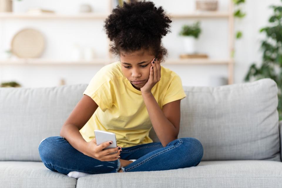 A teenager girl sat on a couch holding her phone and looking sad