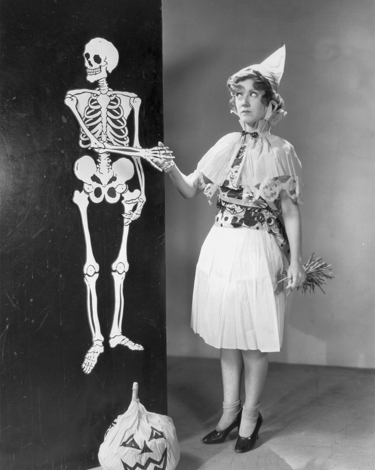 A costumed woman shakes hands with a life-sized Halloween skeleton decoration hanging on a wall.