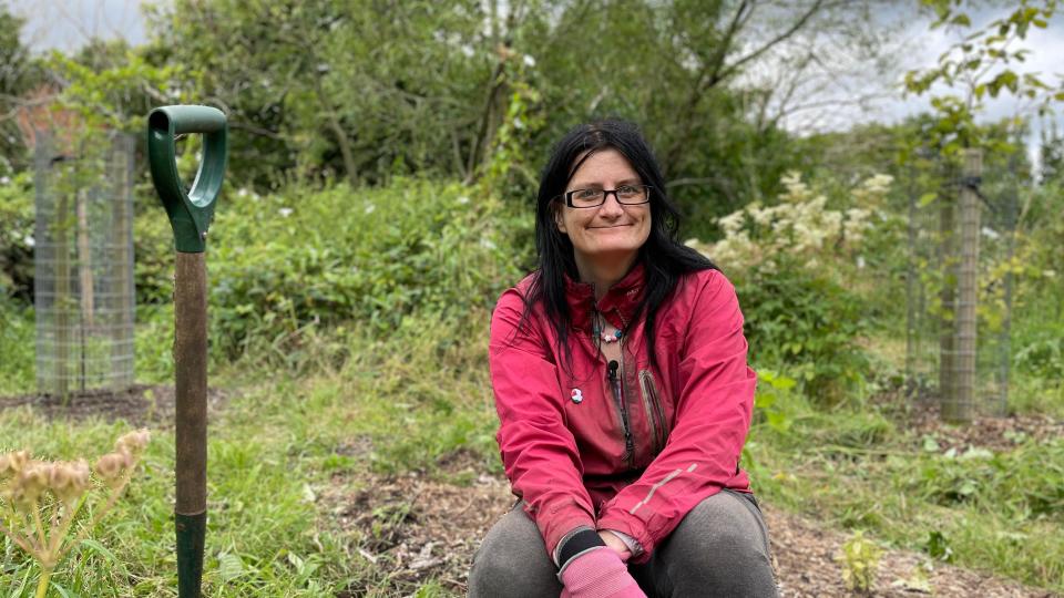 Ruth Mottram at Kingdom Forest Garden. She is crouched by a spade on an area of rough ground with saplings growing behind her