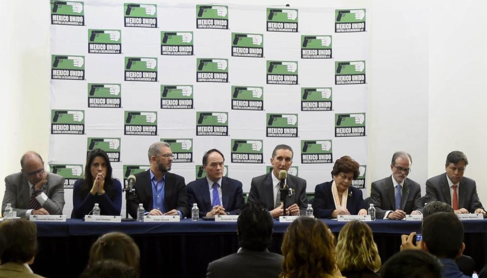 Eight people sit at a long table with microphones; press is visible in the foreground