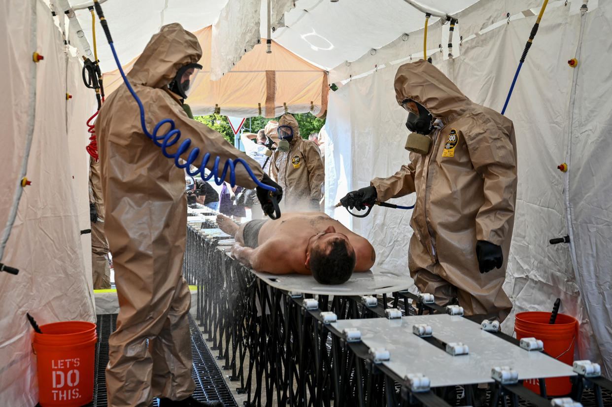 People in full rubber suits and gas masks hold hoses and spray a man lying on his back on a table inside a tent next to an orange bucket labeled: Let's do this.