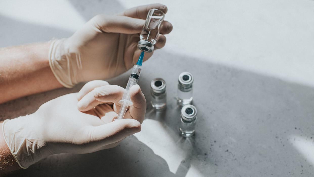  Gloves hands drawing liquid from a vial using a syringe; additional vials are pictured beneath the hands on a table. 