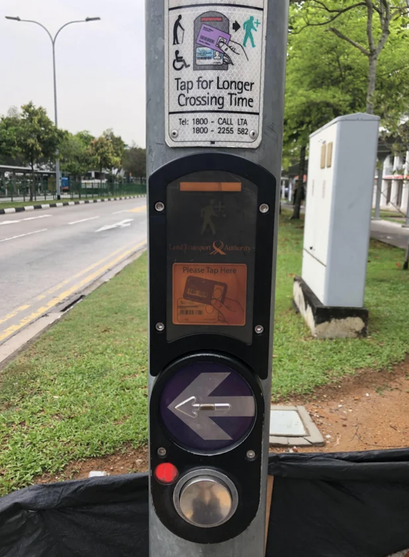 Pedestrian signal button with a sign above instructing to tap for longer crossing time, and an arrow indicating where to tap