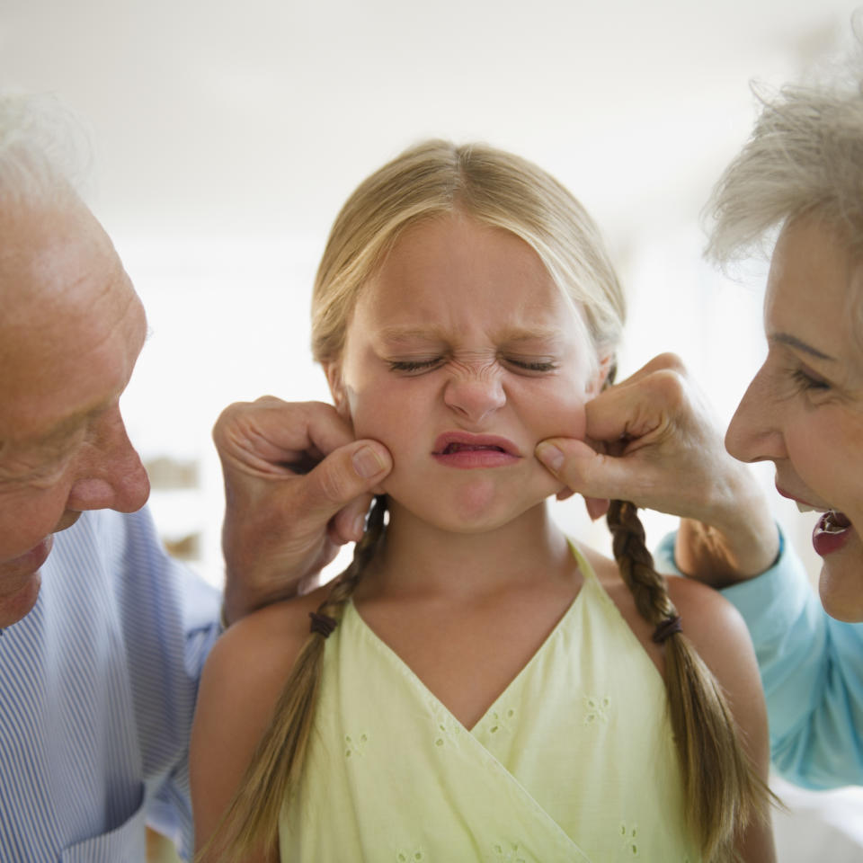 An elderly man and woman gently pinch the cheeks of a young girl with braided hair and a scrunched-up face