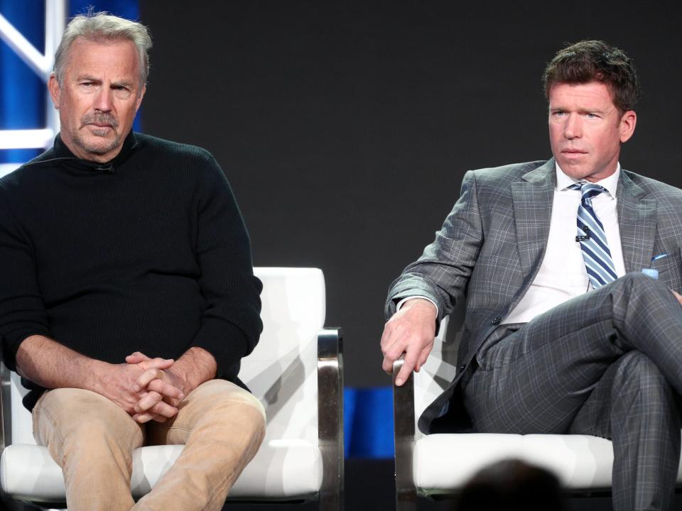 Kevin Costner and Taylor Sheridan sitting in white chairs onstage while listening to someone ask a question.