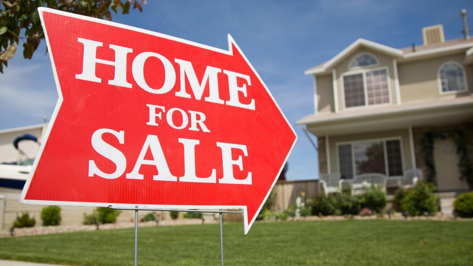 An arrow shaped red "Home For Sale" sign in front of a suburban 2-story home.